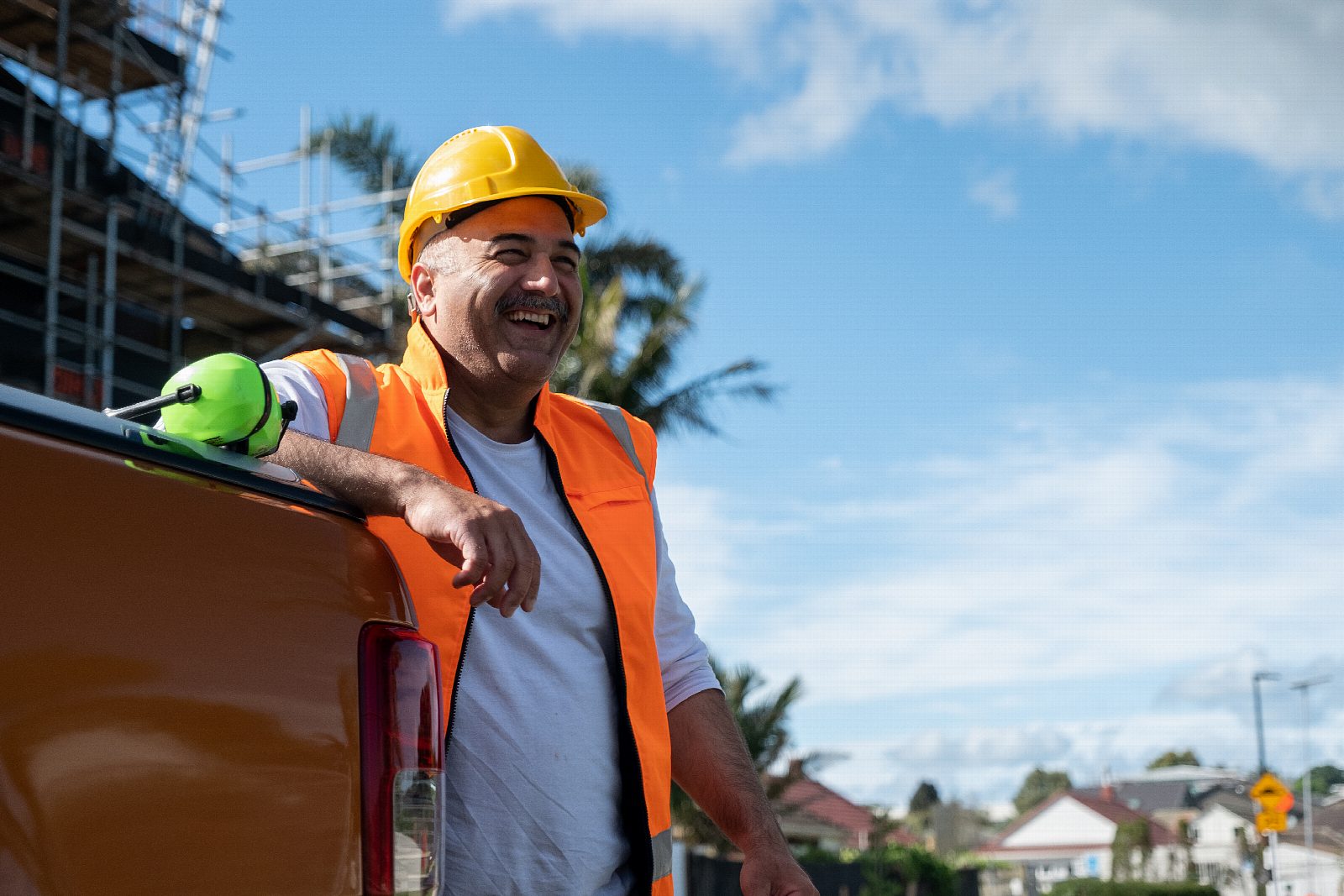 A builder in a hi-vis vest and hard hat leaning on his ute, laughing, with scaffolding behind him